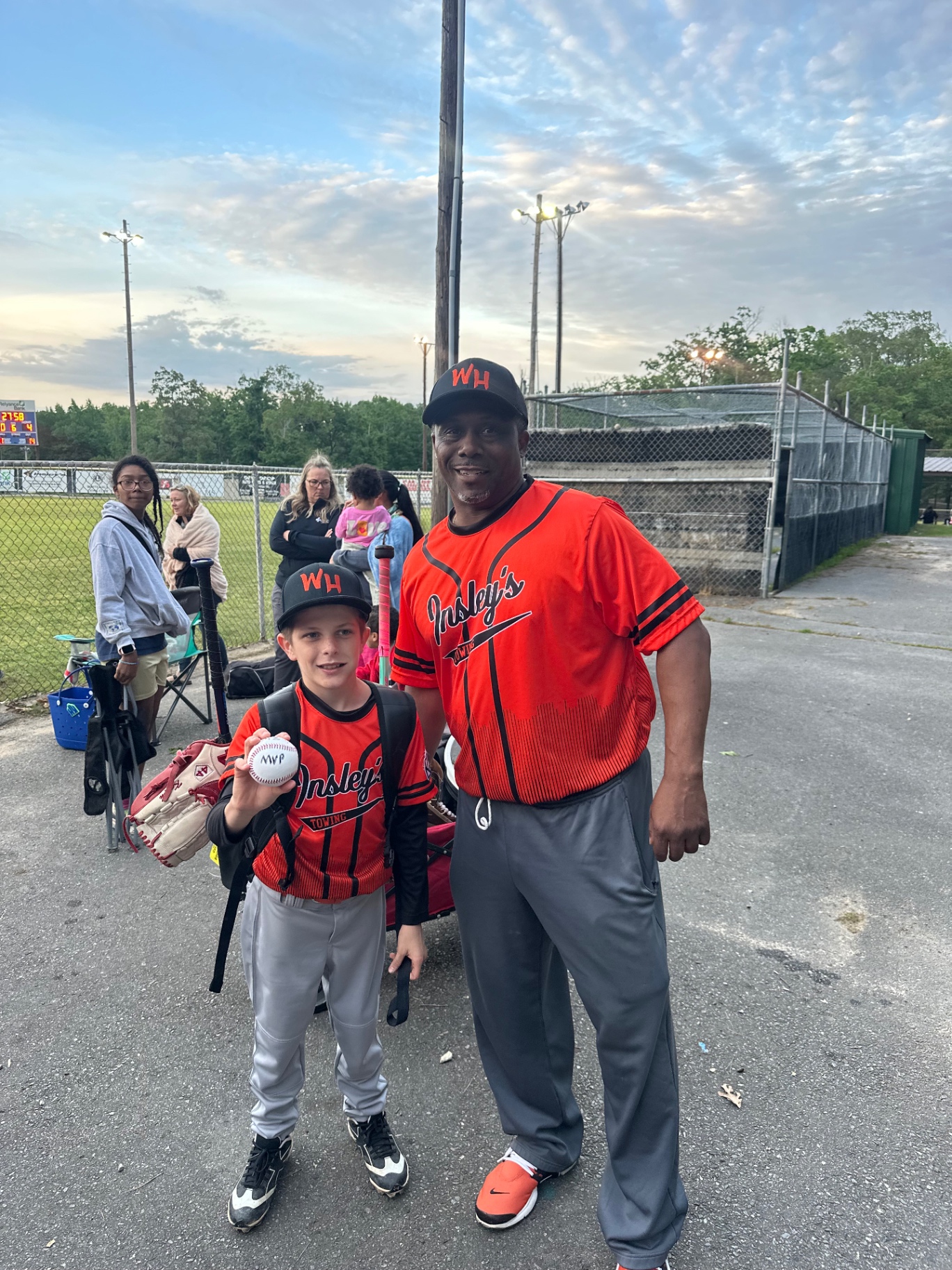 A coach and a young athlete in red jerseys, the athlete holding an MVP baseball at the field