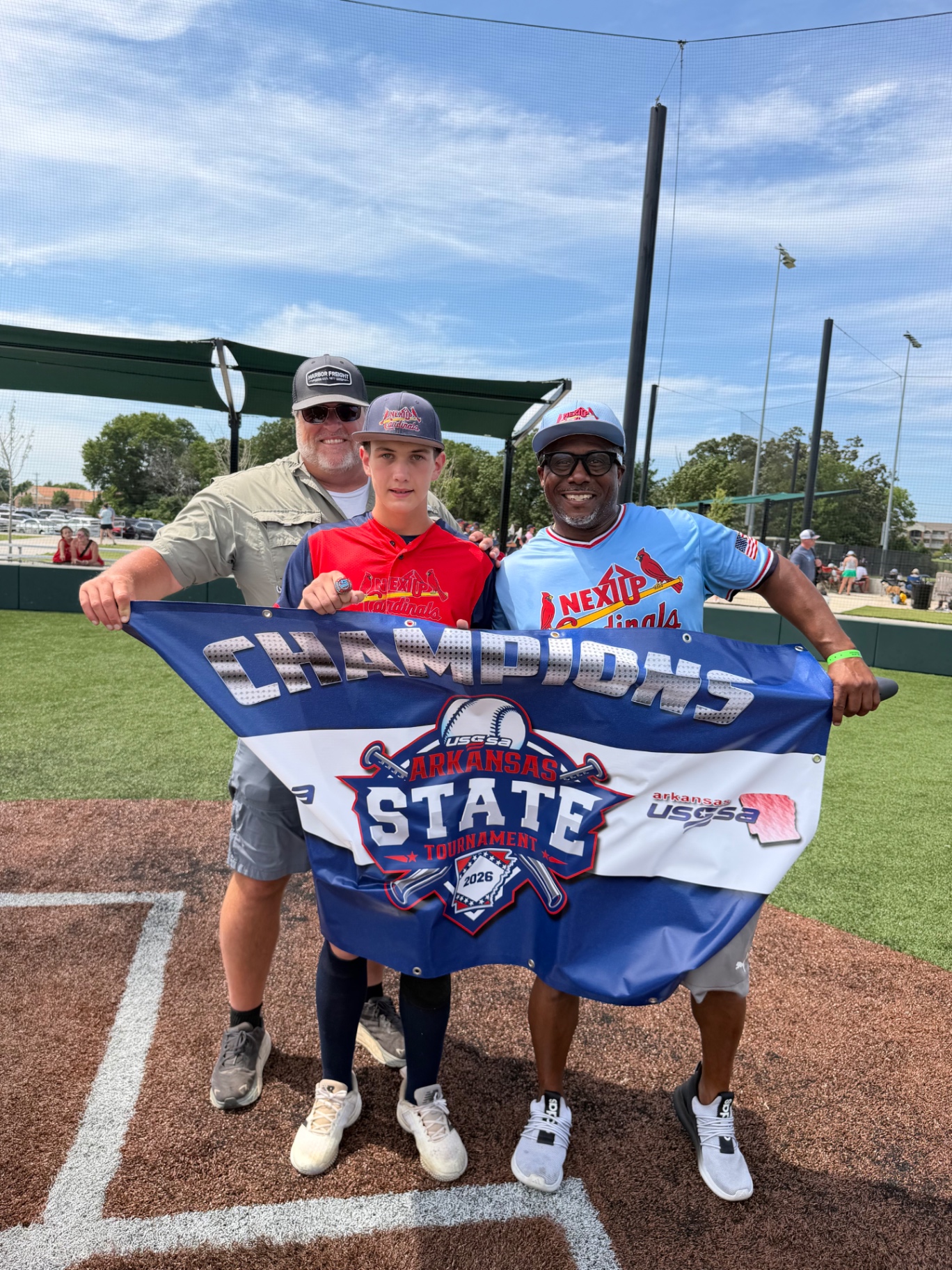 A young baseball player standing with two coaches and holding an Arkansas State Tournament champions banner